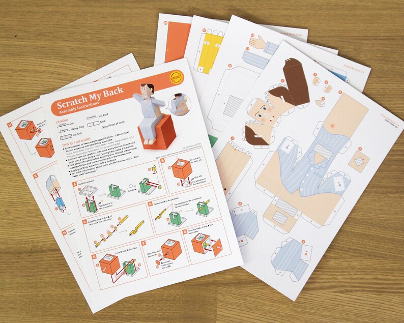 May include: A set of paper craft templates titled Scratch My Back with assembly instructions and printed parts for a character in blue striped pyjamas sitting on an orange cube. The sheets include diagrams, cut lines, and fold markers.