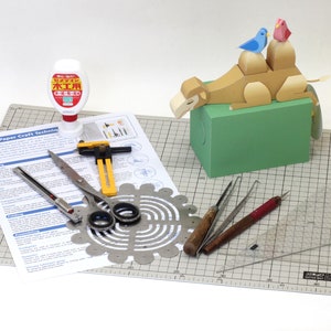 May include: A papercraft camel with two birds on its back, a green base, and a set of papercraft tools including a ruler, scissors, a scoring tool, a cutting tool, a circle cutter, and a bottle of wood glue. The tools are on a gray cutting mat with a grid pattern.