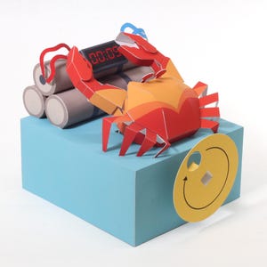 Può includere: Una scultura di carta stravagante con un granchio rosso e arancione appollaiato su un blocco blu. Il granchio interagisce con una bomba, con un timer digitale che mostra "00:09". È presente anche un disco giallo con una faccia sorridente.