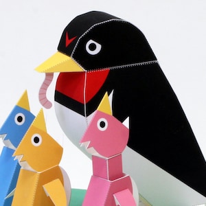 May include: A papercraft model of a black and white bird with a red breast, holding a worm in its beak. Three smaller papercraft birds, one blue, one yellow, and one pink, are standing in front of the larger bird.