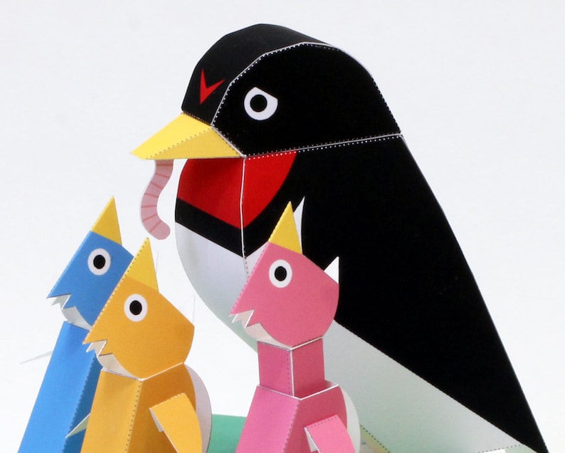 May include: A papercraft model of a black and white bird with a red breast, holding a worm in its beak. Three smaller papercraft birds, one blue, one yellow, and one pink, are standing in front of the larger bird.