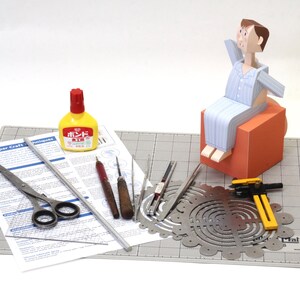 May include: A paper craft kit featuring a paper figure in blue striped pyjamas sitting on an orange cube, accompanied by a yellow glue bottle, metal scissors, a metal ruler, two wooden-handled tools, a metal compass, and a printed instruction sheet.