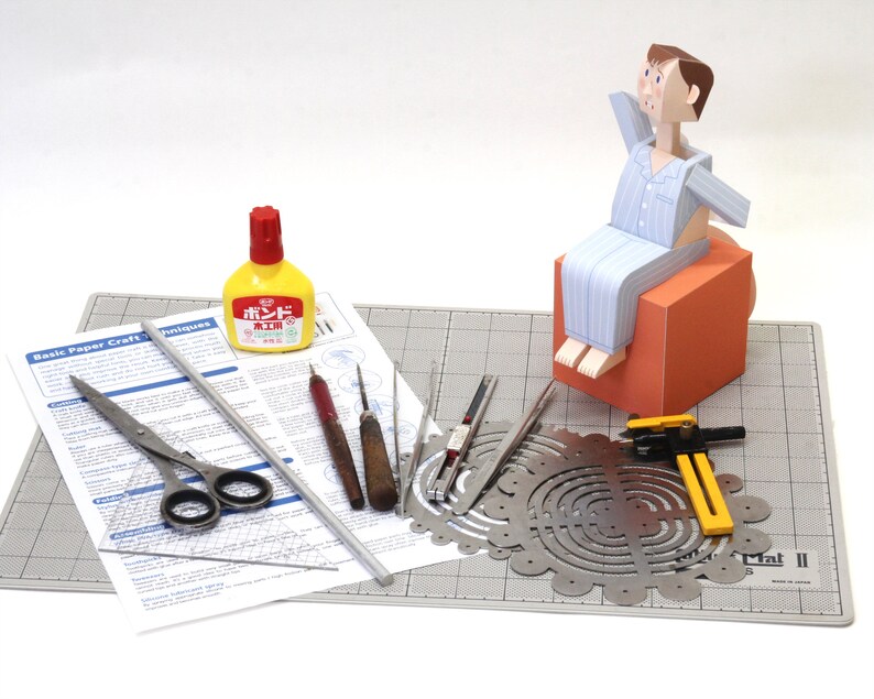 May include: A paper craft kit featuring a paper figure in blue striped pyjamas sitting on an orange cube, accompanied by a yellow glue bottle, metal scissors, a metal ruler, two wooden-handled tools, a metal compass, and a printed instruction sheet.
