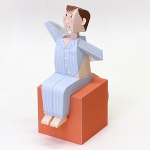 Puede incluir: Una figura de papel de una persona con pijama a rayas azules y blancas sentada en un pequeño cubo naranja.