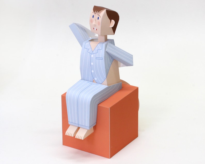May include: A papercraft figure of a person with brown hair wearing light blue and white striped pyjamas, sitting on a solid orange cube. The figure has a surprised facial expression with wide blue eyes and an open mouth, with arms raised.