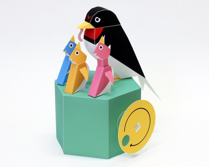 May include: A papercraft model of a black and white bird with a red breast feeding three baby birds. The baby birds are yellow, blue, and pink. The model is on a green hexagonal base with a yellow circular object attached to the side.