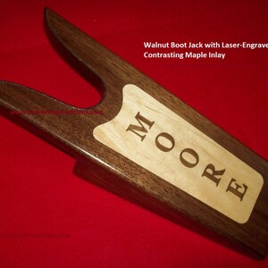 May include: A dark brown walnut boot jack with a laser-engraved maple inlay. The inlay features the name "MOORE". This wooden boot jack is designed to assist in removing boots. The boot jack is a dark brown color.
