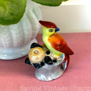 May include: A ceramic bird figurine with red, yellow, and orange paint. The bird is perched on a small, round base with three black and white balls. The base is painted in shades of gray and white.