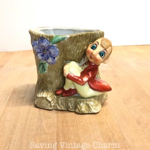 May include: Vintage ceramic planter shaped like a tree trunk with a seated elf figure. The planter features a blue flower and green leaves. The elf wears a red jacket, yellow pants, and red shoes. The planter is a decorative item.