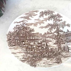 May include: A brown and white oval ceramic plate with a vintage design of a countryside scene. The plate features a brown and white cottage, trees, a horse-drawn carriage, and a river. The plate has a brown rim and a white center.