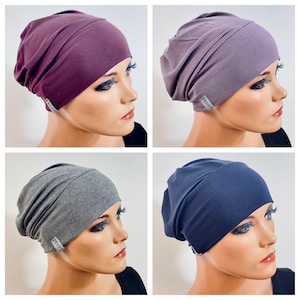 Könnte beinhalten: Eine Kollektion aus vier weichen Beanie-Mützen in Dunkelpflaume, Staublavendel, Graumeliert und Marineblau. Jede Mütze aus weichem Stoff hat ein breites Band und ein kleines graues Etikett an der Seite mit dem Text "I LOVE KOPY" in schwarzer Schrift.