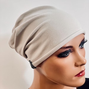 Könnte beinhalten: Eine hellbeige Beanie-Mütze aus weichem Stoff. Die Mütze wird auf einem Schaufensterkopf gezeigt. Die Mütze hat ein kleines schwarzes Etikett an der Seite. Die Mütze ist neutral und kann von jedem getragen werden.