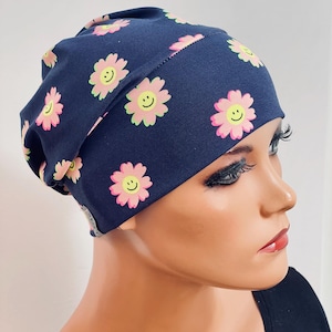 Könnte beinhalten: Eine marineblaue Beanie-Mütze mit einem sich wiederholenden Muster aus rosa und gelben lächelnden Gänseblümchen. Die Mütze ist aus einem weichen Stoff gefertigt und ist zum Tragen auf dem Kopf bestimmt.