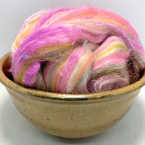 May include: A bowl filled with colorful wool roving. The roving is a blend of pink, yellow, and white fibers. The bowl is brown with a speckled glaze.