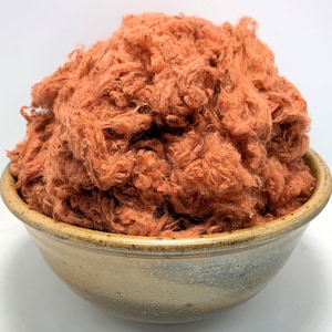 May include: A bowl filled with a large amount of loose, orange-colored fiber. The fiber is fluffy and appears to be made of natural materials.