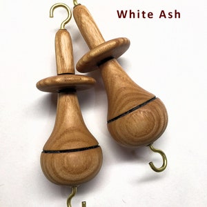 May include: Two wooden spinning whorls with brass hooks. The whorls are made of white ash wood and have a teardrop shape. The text "White Ash" is visible in the image.