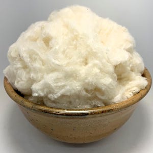 May include: A mound of fluffy, off-white raw cotton fibers piled high inside a round, speckled tan ceramic bowl. The bowl features a smooth, glazed finish with darker brown flecks and a slightly darker rim, contrasting with the soft, fibrous texture.
