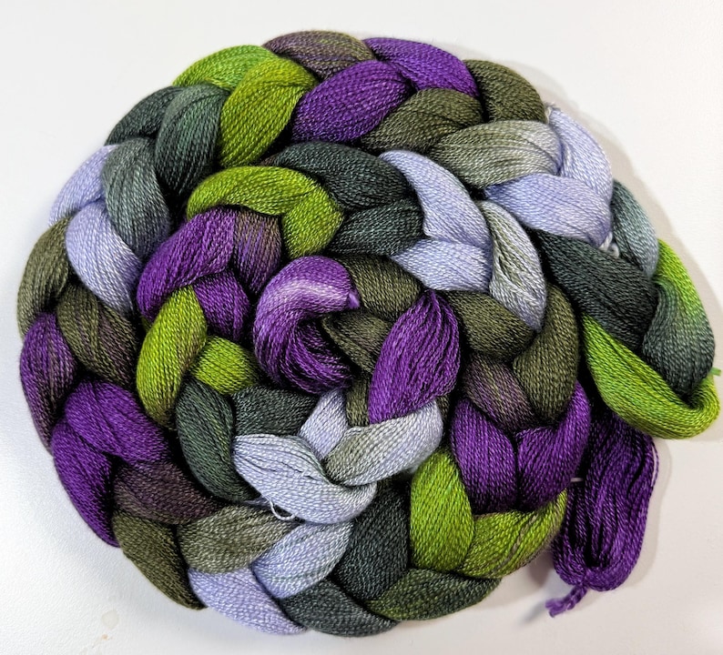 May include: A circular braid of colorful yarn in shades of green, purple, and gray. The yarn is tightly woven, creating a textured surface. The colors blend together, creating a visually appealing pattern. The yarn is likely for knitting or crocheting projects.