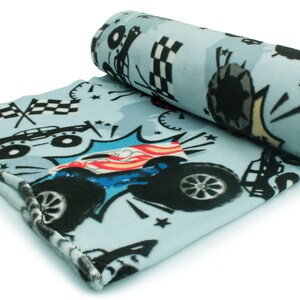 May include: A blue fleece blanket with a monster truck design. The blanket features a black monster truck with red, white, and blue accents, and a checkered flag pattern.