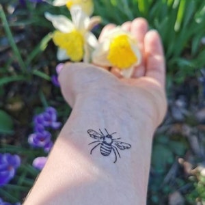 Può includere: Un polso con un piccolo tatuaggio di ape in bianco e nero. La persona tiene un mazzo di narcisi gialli in mano.