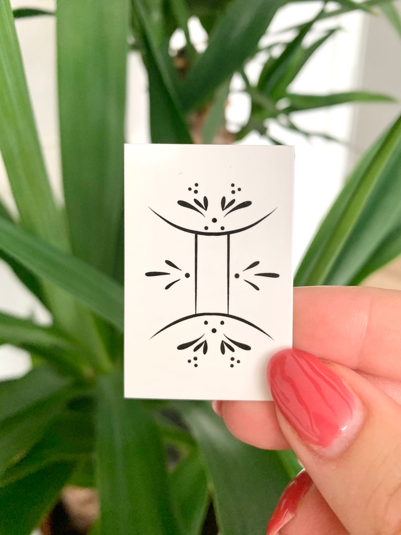 May include: A white square temporary tattoo with a black outline of the Gemini zodiac symbol. The symbol is stylized with a simple design of two connected circles with small dots and lines.