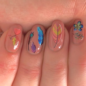 Op de afbeelding: Close-up van een hand met vijf vingernagels die in een licht perzikkleurige kleur zijn geverfd. Elke nagel heeft een ander ontwerp met een hart, een veer, een pauwenveer, een bloem en een veer met stippen.