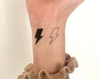 May include: A person's wrist with two lightning bolt tattoos. One tattoo is black and filled in, the other is an outline.