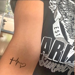 May include: A black temporary tattoo on a person's arm. The tattoo features a cross, a heartbeat line, and a heart.
