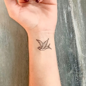 Pode incluir: Um desenho de linha simples de uma tatuagem de andorinha no pulso de uma pessoa.