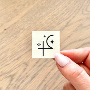 May include: A white square sticker with a black crescent moon, a cross, and three stars. The sticker is a celestial design.