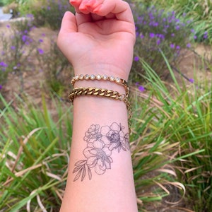 May include: A close-up of a person's arm with a black floral tattoo on their wrist. The tattoo features a cluster of flowers with delicate stems and leaves. The person is wearing two gold bracelets, one with a chain design and the other with small clear stones.