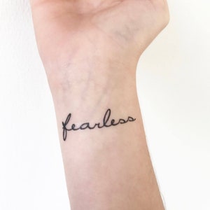 May include: A close-up of a person's wrist with a simple black tattoo that reads "fearless" in cursive script.