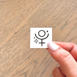 Op de afbeelding: Een witte vierkante sticker met een zwart astrologisch symbool voor de planeet Venus. Het symbool is een cirkel met een kruis eronder en een halvemaanvorm boven de cirkel. Er zijn twee kleine sterren aan weerszijden van het symbool.