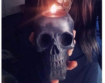 May include: A black skull-shaped candle holder with a lit candle inside. The candle holder is being held by a person wearing a black beanie, black glasses, and a black lace top.