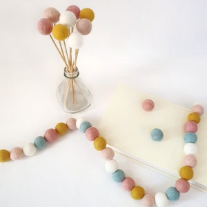 Bloom pastel pom pom garland made from wool felt balls, in the colours Ivory, Pastel Blue, Rose, Mustard, Blush. Garland shown lying flat across a table with felt ball flowers in a vase, in the background.