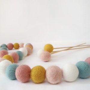 Bloom pastel pom pom garland made from wool felt balls, in the colours Ivory, Pastel Blue, Rose, Mustard, Blush. Garland shown lying flat across a table with felt ball flowers in the background.