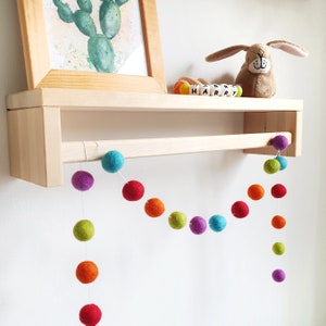 Pom Pom Felt Ball Garland shown hanging from a shelf, in Rainbow colours