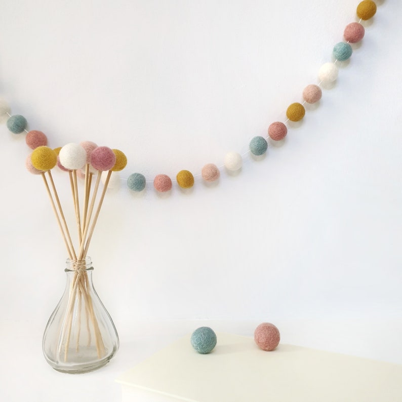 Bloom pastel pom pom garland made from wool felt balls, in the colours Ivory, Pastel Blue, Rose, Mustard, Blush. Garland shown hanging across the wall with felt ball flowers in a vase.