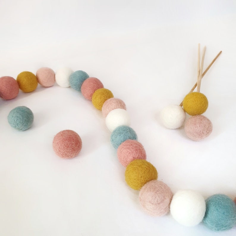 Bloom pastel pom pom garland made from wool felt balls, in the colours Ivory, Pastel Blue, Rose, Mustard, Blush. Garland shown lying flat across a table with felt ball flowers in the background.