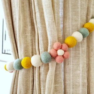 May include: A curtain tieback made of a string of felt balls in yellow, white, and light blue colors. The string ends with a pink felt flower with a white center.