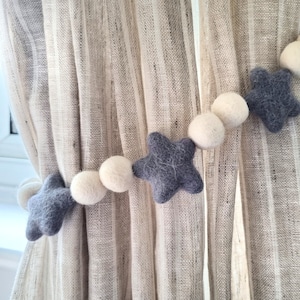 May include: A gray and white felt garland with stars and pom poms. The garland is hanging on a beige curtain.