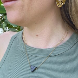May include: A gold chain necklace with a black triangular lava rock pendant. The necklace is layered over a second gold chain. The person is wearing a green tank top and gold earrings with a brown leaf design.
