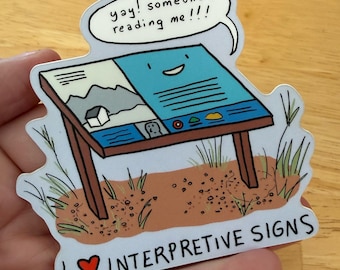May include: A die-cut sticker featuring an illustrated interpretive sign on a brown dirt patch with green grass. The sign has a blue face with a smiling expression and a speech bubble that reads, "yay! someone's reading me!!!". Text below reads, "I ❤️ INTERPRETIVE SIGNS".
