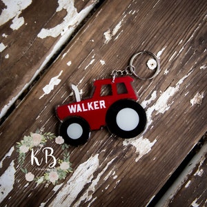 Può includere: Portachiavi a forma di trattore rosso con il nome "WALKER" in bianco. Il portachiavi ha ruote nere e bianche e un portachiavi argentato. Sfondo in legno invecchiato.