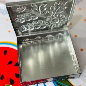 May include: A small, open, silver-colored metal box with a hinged lid. The lid features an embossed floral design, including a sunflower and leaves. The box is sitting on a colorful tablecloth with a watermelon design.