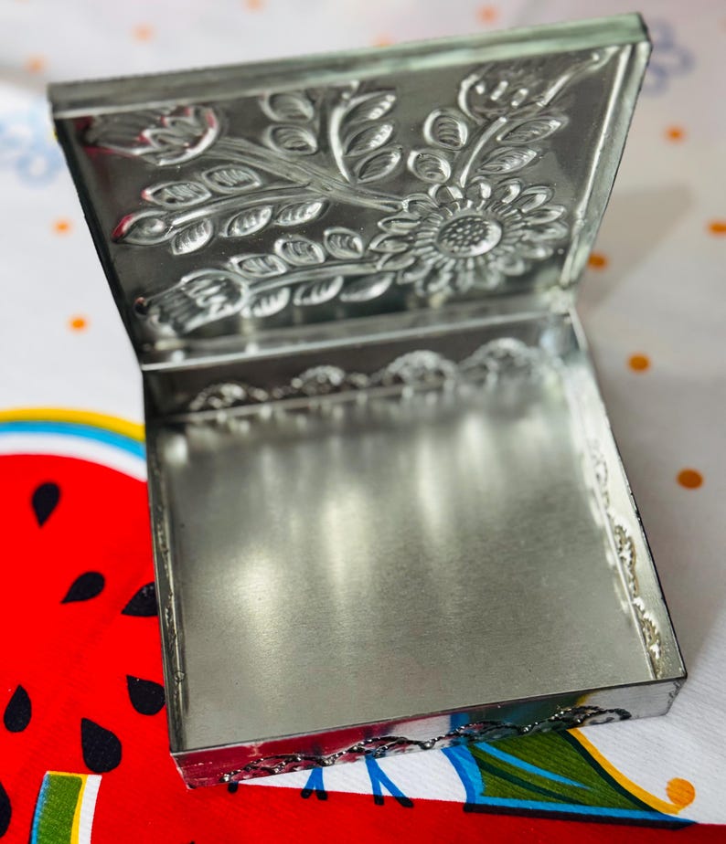 May include: A small, open, silver-colored metal box with a hinged lid. The lid features an embossed floral design, including a sunflower and leaves. The box is sitting on a colorful tablecloth with a watermelon design.