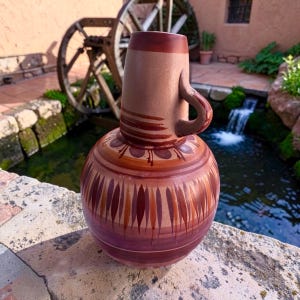 May include: A terracotta pitcher with a rounded body and a single handle. The vessel features horizontal bands and vertical leaf-like patterns in shades of dark brown, reddish-brown, and tan, with a smooth, matte finish on the neck and body.