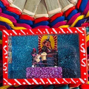 May include: A rectangular, red-framed diorama featuring a wrestling ring with two figures, set against a blue glitter background. The diorama is placed on a colorful woven base with red, blue, yellow, and green ribbons.