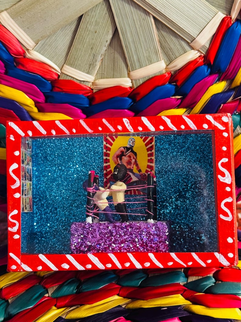 May include: A rectangular, red-framed diorama featuring a wrestling ring with two figures, set against a blue glitter background. The diorama is placed on a colorful woven base with red, blue, yellow, and green ribbons.