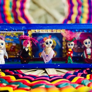 May include: A blue framed display case featuring five Day of the Dead figurines. Each figurine has a skull face and is dressed in colorful attire. The background is a colorful, woven pattern.